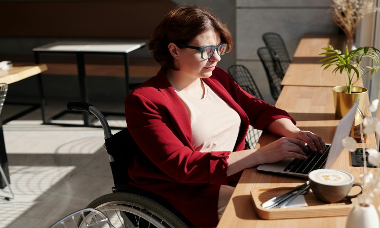 Woman in wheelchair sitting in a cafe working on a laptop.